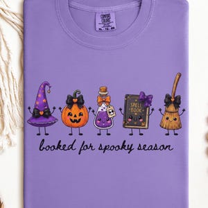 A folded  violet Comfort Colors t-shirt featuring a graphic of five cartoon characters: a witch hat, a jack-o'-lantern, a potion bottle, a spell book, and a broom. The text below the characters reads "booked for spooky season" in a black cursive font.