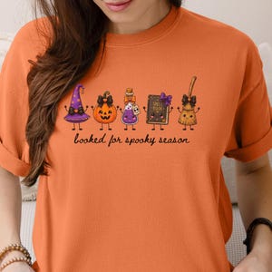 A model wearing a melon orange Comfort Colors tee shirt featuring a graphic of five cartoon characters: a purple witch hat, a jack-o'-lantern, a potion bottle, a spell book, and a broomstick, all with bows. Below the characters is the text, booked for spooky season.