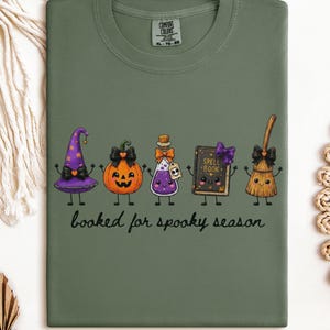 A folded sage green Comfort Colors t-shirt featuring a graphic of five cute, anthropomorphic Halloween items: a purple witch hat, a jack-o'-lantern, a purple potion bottle, a spell book, and a broom. The text below reads, "booked for spooky season."