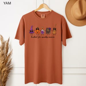 A hangimg yam orange Comfort Colors short-sleeve tee featuring a graphic of a purple witch hat, an orange jack-o'-lantern, a potion bottle, a spell book, and a broomstick with the text "booked for spooky season" printed in black cursive lettering.