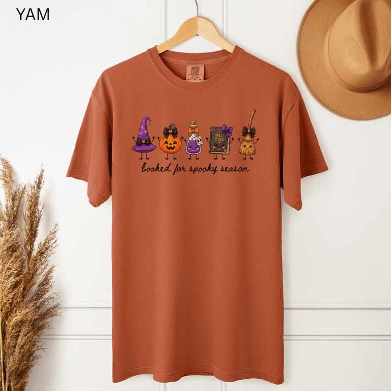 A hangimg yam orange Comfort Colors short-sleeve tee featuring a graphic of a purple witch hat, an orange jack-o'-lantern, a potion bottle, a spell book, and a broomstick with the text "booked for spooky season" printed in black cursive lettering.
