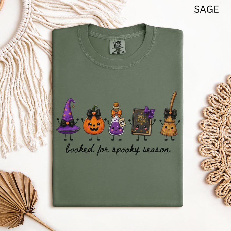 A folded sage green Comfort Colors t-shirt featuring a graphic of five cute, anthropomorphic Halloween items: a purple witch hat, a jack-o'-lantern, a purple potion bottle, a spell book, and a broom. The text below reads, "booked for spooky season."