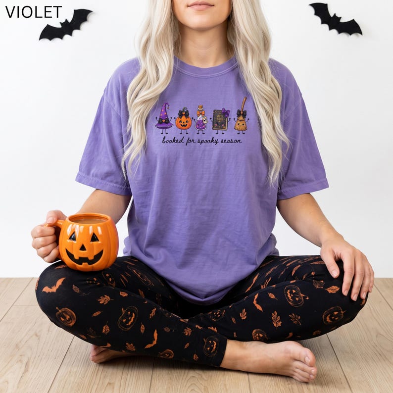 A model wearing a violet Comfort Colors  short-sleeve Halloween t-shirt featuring a graphic of a witch hat, pumpkin, potion bottle, book, and broom with the text "booked for spooky season". The shirt is paired with black leggings patterned with orange pumpkins and bats.