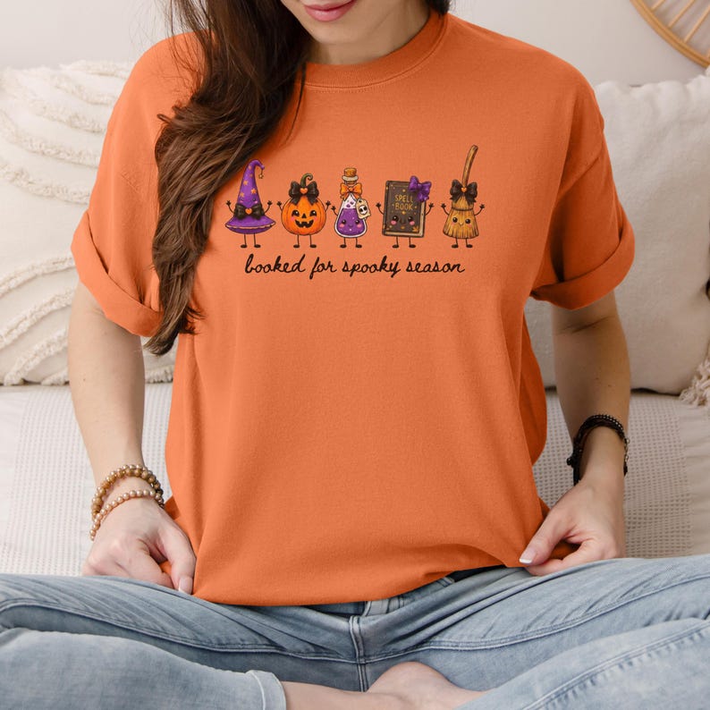 A model wearing a melon orange Comfort Colors tee shirt featuring a graphic of five cartoon characters: a purple witch hat, a jack-o'-lantern, a potion bottle, a spell book, and a broomstick, all with bows. Below the characters is the text, booked for spooky season.