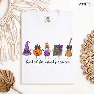 A folded white Comfort Colors tee featuring a graphic of five cute, anthropomorphic Halloween items: a purple witch hat, an orange jack-o'-lantern, a purple potion bottle, a spell book, and a broom, with the text "booked for spooky season" below.