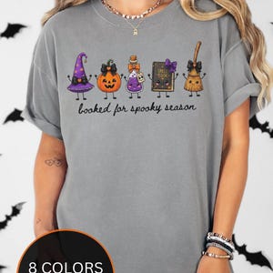 A model wearing a grey Comfort Colors t-shirt featuring a graphic of a purple witch hat, a jack-o'-lantern, a potion bottle, a spell book, and a broomstick with the text "booked for spooky season" printed in black cursive script below the illustrated characters.