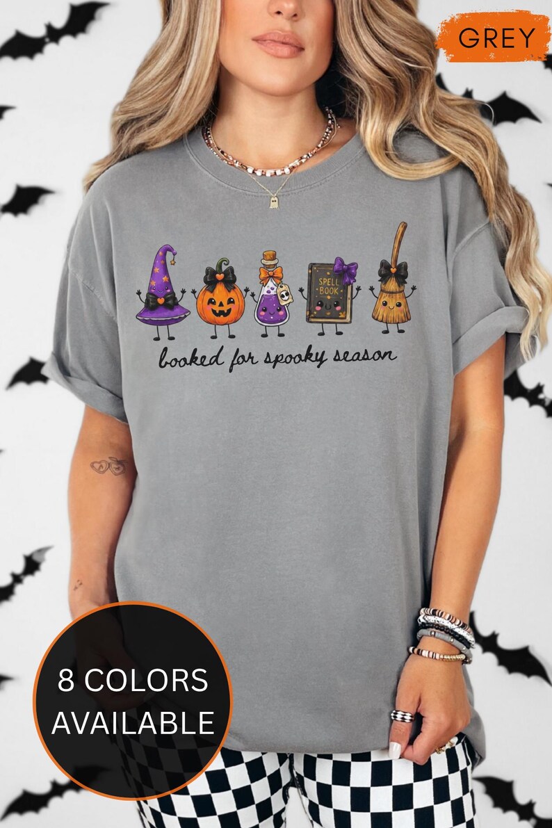 A model wearing a grey Comfort Colors t-shirt featuring a graphic of a purple witch hat, a jack-o'-lantern, a potion bottle, a spell book, and a broomstick with the text "booked for spooky season" printed in black cursive script below the illustrated characters.