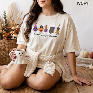 A model wearing an ivory Comfort Colors t-shirt featuring a graphic of a purple witch hat, a jack-o'-lantern, a potion bottle, a spell book, and a broomstick with the text "booked for spooky season" printed in black cursive script across the chest area.
