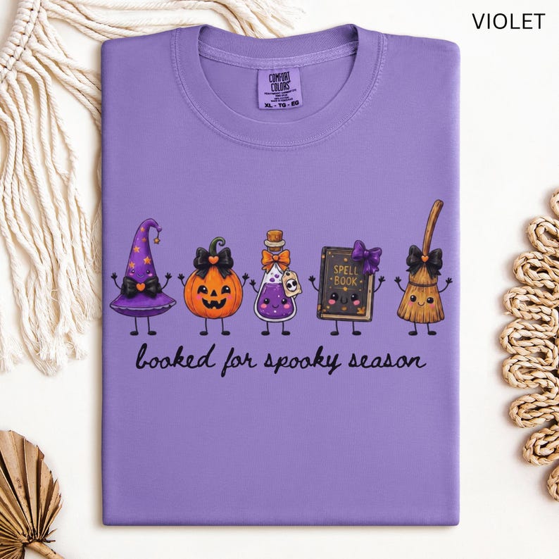 A folded  violet Comfort Colors t-shirt featuring a graphic of five cartoon characters: a witch hat, a jack-o'-lantern, a potion bottle, a spell book, and a broom. The text below the characters reads "booked for spooky season" in a black cursive font.