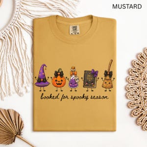 A folded mustard yellow Comfort Colors t-shirt featuring a graphic of five cartoon characters: a witch hat, a jack-o'-lantern, a potion bottle, a spell book, and a broom. The text below the characters reads "booked for spooky season" in a black cursive font.