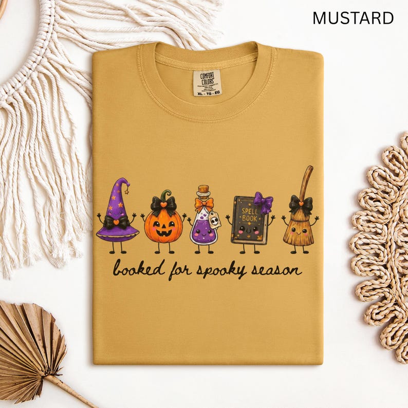 A folded mustard yellow Comfort Colors t-shirt featuring a graphic of five cartoon characters: a witch hat, a jack-o'-lantern, a potion bottle, a spell book, and a broom. The text below the characters reads "booked for spooky season" in a black cursive font.