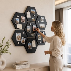 May include: A wall-mounted display of black hexagonal magnetic boards, arranged in a honeycomb pattern. Photos and notes are attached with silver magnets. A person is shown placing a photo on one of the boards. The display is mounted on a white wall.
