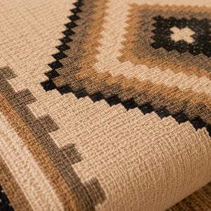 Op de afbeelding: Een geweven textielstof met een geometrisch patroon van herhalende ruitvormen en getrapte randen. Het ontwerp bevat een neutrale beige basiskleur geaccentueerd met tinten bruin, tan en zwart in een gestructureerd, rasterachtig weefsel.