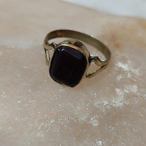 Könnte beinhalten: Ein Vintage-Ring in Goldoptik mit einem dunkelburgunderfarbenen, rechteckigen Edelstein. Der Ring hat ein schlichtes Design mit dekorativen Aussparungen an den Seiten, das einen klassischen, eleganten Stil zeigt.