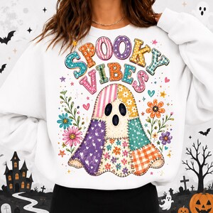 May include: A white long-sleeve sweatshirt featuring a colorful graphic of a ghost made of various patterned fabric patches. The ghost is surrounded by floral illustrations and the text SPOOKY VIBES in multi-colored, patterned letters.