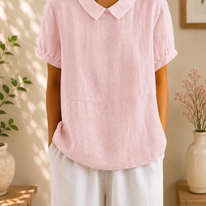 May include: A pale pink linen top with a rounded collar and short sleeves. The top is worn with white linen trousers. The outfit is simple and elegant, suitable for casual wear.