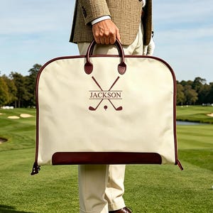 May include: A cream-coloured garment bag with dark maroon trim and handles. The front features a maroon graphic of two crossed golf clubs and a golf ball, with the name JACKSON printed in a horizontal line across the centre of the clubs.