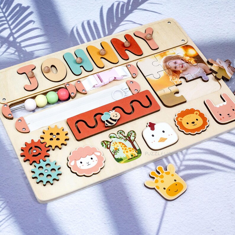 May include: Wooden activity board with the name "JOHNNY" in colorful letters. Features include a zipper, bead maze, puzzle with a photo, and animal-shaped pieces like a lion, sheep, and giraffe. The board is designed for early childhood development.
