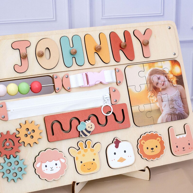 May include: Wooden activity board with the name "JOHNNY" in colorful letters. Features include a zipper, buckle, bead maze, gears, and animal-shaped puzzle pieces. A photo puzzle is also included.