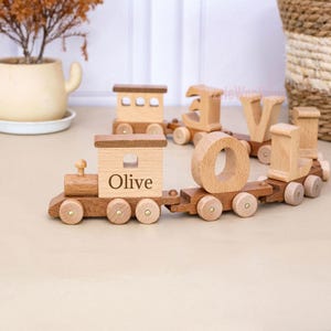 May include: A wooden toy train set featuring a locomotive engine with the name Olive printed on the side, followed by several connected train cars, each carrying a large wooden letter to spell out a name, all with a natural light wood finish.