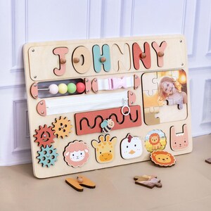 May include: Wooden activity board with the name "JOHNNY" in colorful letters. Features include a zipper, buckle, bead maze, gears, animal shapes, and a photo puzzle. The board is designed for early childhood development and learning.