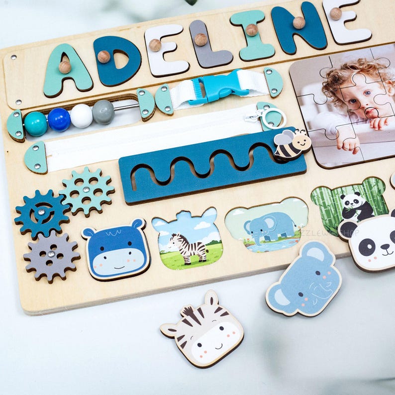 May include: Wooden activity board with the name "ADELINE" in colorful letters. Features include a zipper, buckle, gears, beads, and animal-shaped puzzle pieces. The board has a light wood finish and includes a photo puzzle.