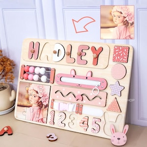 May include: A wooden activity board featuring the name HADLEY in pink and white letters, a bead abacus, a zip, a buckle, and a photo puzzle of a child. The board includes cutouts for numbers 1 through 5 and various shapes like a star and circle.