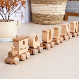May include: A wooden toy train set featuring individual cars that carry the letters Q, U, I, N, N. The engine car is engraved with the name Quinn. Each car has small wheels and is crafted from light and medium-toned wood, arranged in a line.