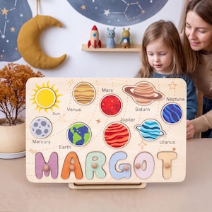 Op de afbeelding: Een rechthoekig houten puzzelbord met een zonnestelselthema, gelabelde planeten en de naam MARGOT in grote, kleurrijke uitgesneden letters. Het bord bevat illustraties van de zon, Mercurius, Venus, Aarde, Mars, Jupiter, Saturnus, Uranus en Neptunus.