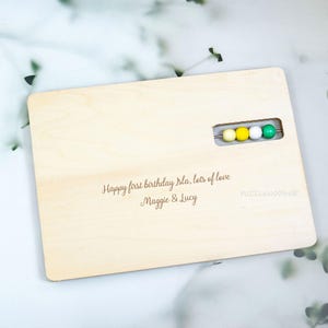May include: A light-colored wooden abacus with rounded corners. The abacus has a row of four colored beads: yellow, white, and green. The text "Happy first birthday Isla, lots of love Maggie & Lucy" is engraved on the wood.