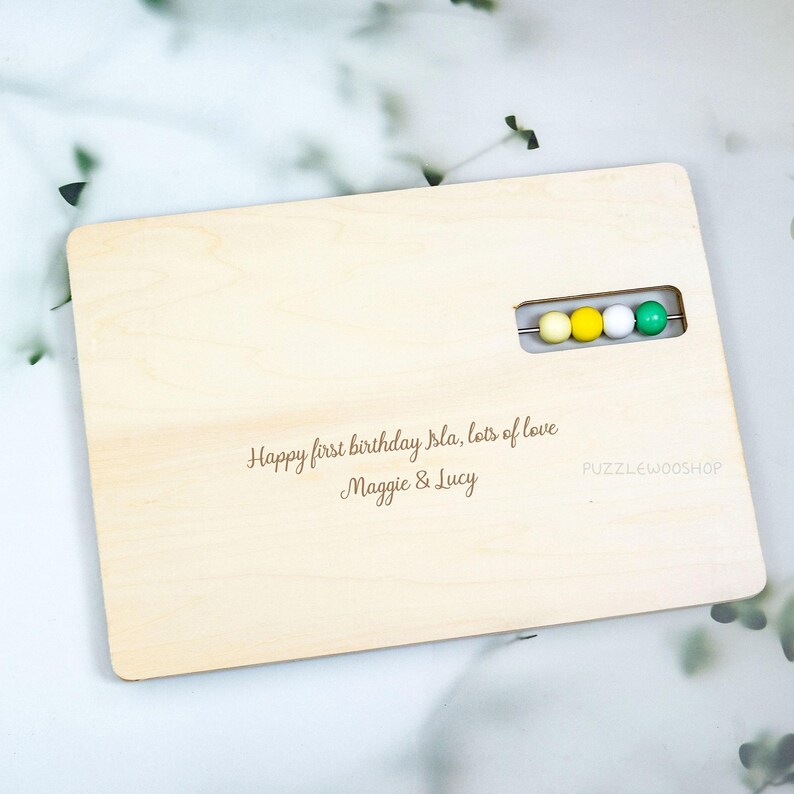May include: A light-colored wooden abacus with rounded corners. The abacus has a row of four colored beads: yellow, white, and green. The text "Happy first birthday Isla, lots of love Maggie & Lucy" is engraved on the wood.