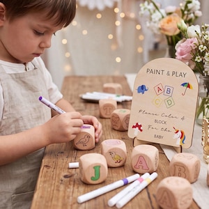 May include: A set of light-coloured wooden blocks featuring engraved letters and illustrations, accompanied by a wooden sign that reads PAINT & PLAY Please Color A BLOCK For the BABY, along with several colourful markers for decorating the blocks.