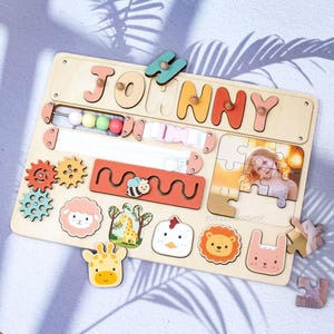 May include: Wooden activity board with the name "JOHNNY" in colorful letters. Features include gears, a bead maze, a buckle, a puzzle with a photo, and animal shapes like a giraffe, sheep, and lion. The board is designed for early childhood development.