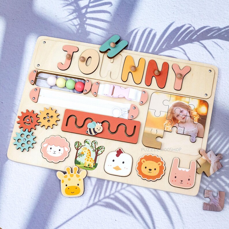May include: Wooden activity board with the name "JOHNNY" in colorful letters. Features include gears, a bead maze, a buckle, a puzzle with a photo, and animal shapes like a giraffe, sheep, and lion. The board is designed for early childhood development.