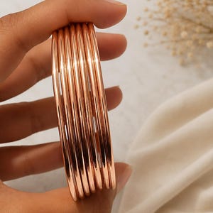 May include: A set of seven thin, polished rose gold-colored bangles stacked together. Each bangle features a smooth, rounded, and reflective metallic surface. The bracelets are uniform in thickness and circular in shape, creating a cohesive set.