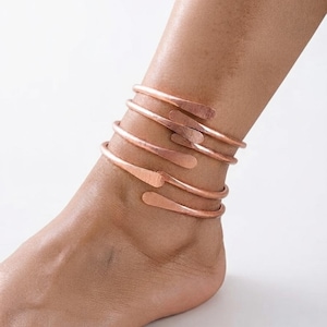 May include: A copper anklet featuring a multi-coil wrap design with a hammered texture. The metal band spirals around the ankle, ending in rounded, flattened tips. The surface of the copper has a warm, metallic reddish-brown finish.