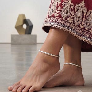 May include: A pair of smooth, polished silver anklets worn around the ankles. The anklets are simple, circular bands with a reflective metallic finish. The jewelry is paired with a deep maroon fabric featuring intricate light-colored embroidery.