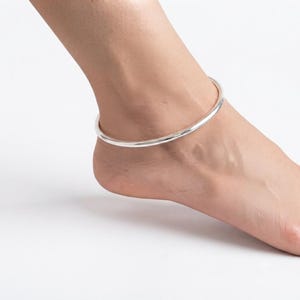 May include: A simple, polished silver-tone anklet featuring a smooth, continuous circular band design. The metallic accessory rests around the ankle, showcasing a reflective, high-shine finish that catches the light with its clean, minimalist aesthetic.