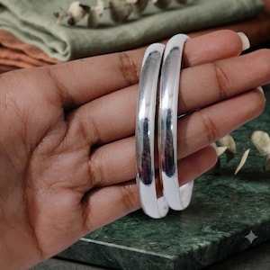 May include: Two smooth, polished silver-toned bangles with a rounded, high-shine surface. These circular bracelets feature a simple, solid design with a reflective metallic finish that catches the light, creating a clean and uniform appearance.