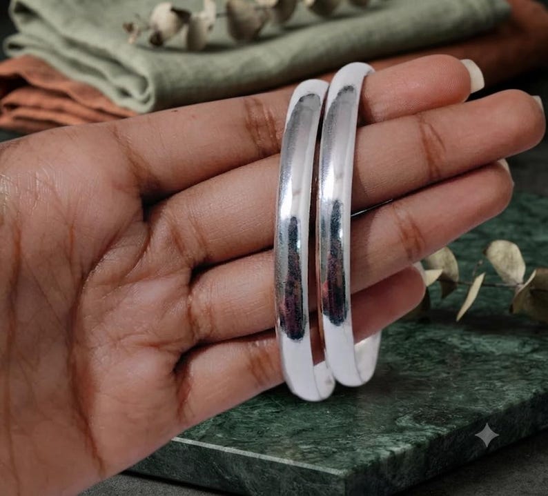 May include: Two smooth, polished silver-toned bangles with a rounded, high-shine surface. These circular bracelets feature a simple, solid design with a reflective metallic finish that catches the light, creating a clean and uniform appearance.