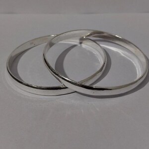 May include: Two polished silver-toned bangle bracelets with a smooth, rounded surface. The two circular bangles are interlocked, creating a continuous metallic loop. The reflective surface of the metal catches the light, highlighting the simple, clean design of the jewelry pieces.