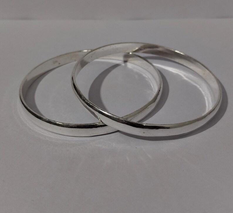 May include: Two polished silver-toned bangle bracelets with a smooth, rounded surface. The two circular bangles are interlocked, creating a continuous metallic loop. The reflective surface of the metal catches the light, highlighting the simple, clean design of the jewelry pieces.