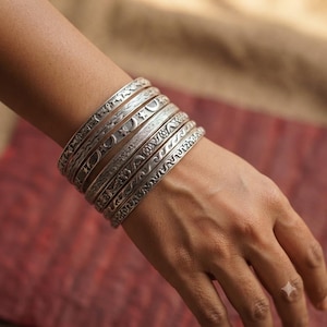 Peut inclure: Un empilement de six bracelets rigides en métal argenté portés au poignet. Chaque bracelet présente un motif en relief distinct, incluant des symboles célestes comme des étoiles et des croissants de lune, des lignes ondulées et des motifs floraux.