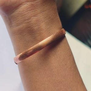 May include: A solid copper bangle bracelet with a smooth, polished finish. The bracelet features a simple, continuous circular band design with a warm, metallic reddish-brown hue that reflects light across its rounded surface.