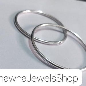 May include: Two polished silver-toned circular hoop earrings with a smooth, reflective finish. The hoops are interlocked, showcasing a simple, thin, and rounded metal design. The text BhawnaJewelsShop is printed in black at the bottom.