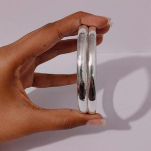 May include: Two polished silver-tone bangles with a smooth, rounded surface. The bracelets feature a reflective, metallic finish and a simple, circular design. These two identical bangles are held together, showcasing their uniform shape and shine.