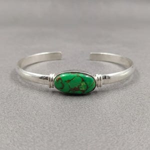 May include: A silver cuff bracelet featuring a central oval-shaped green turquoise stone with brown matrix veining. The metal band has a smooth, polished finish and includes decorative ribbed detailing on both sides of the stone setting.