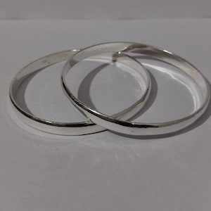 May include: Two polished silver-toned bangle bracelets with a smooth, rounded surface. The two circular bangles are interlocked, creating a continuous metallic loop. The reflective surface of the metal catches the light, highlighting the simple, clean design of the jewelry pieces.