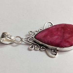 May include: A teardrop-shaped pendant featuring a faceted, deep pink gemstone set in a silver-toned metal frame. The setting includes intricate, swirling wirework details and a decorative bail at the top for attaching to a chain.