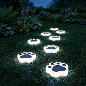 May include: A series of solar-powered outdoor path lights shaped like animal paw prints. Each light features a glowing white border and a dark, rectangular solar panel in the center, arranged in a trail across a dark green grassy lawn.